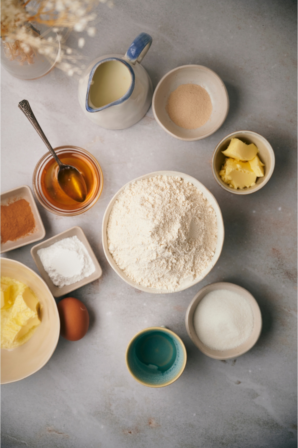 An assortment of ingredients including bowls of flour, butter, sugar, yeast, honey, powdered sugar, an egg and a container of milk.