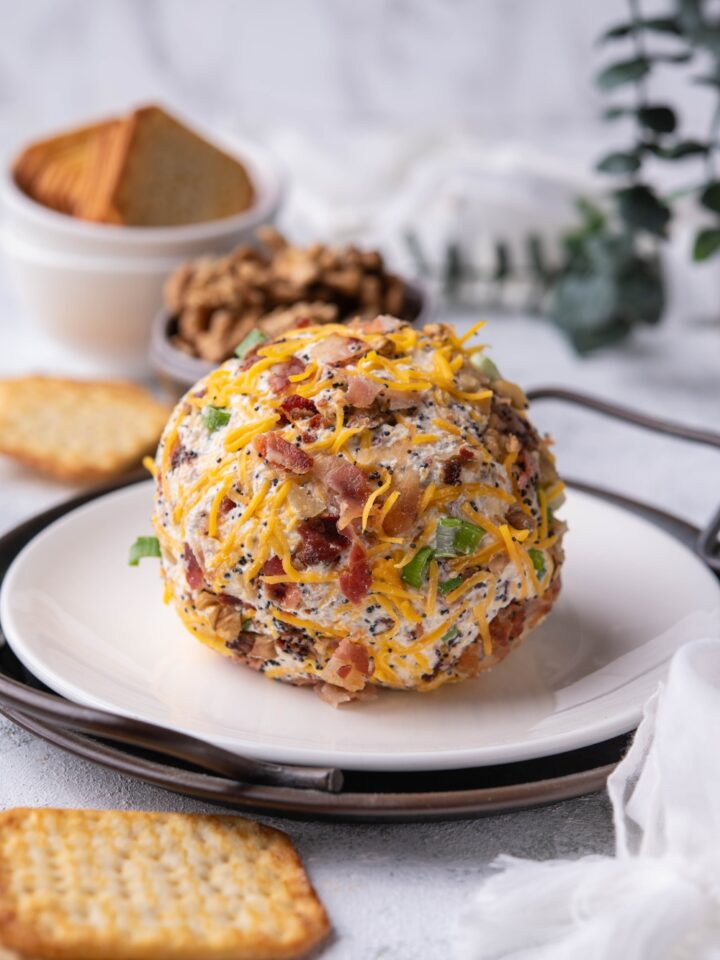 A cheese ball on a white plate layered on a round serving dish, surrounded by crackers and a bowl of chopped nuts.