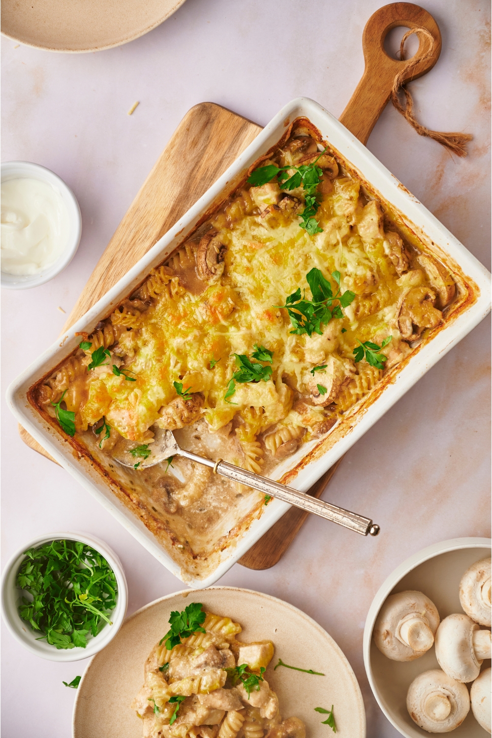 Chicken and mushroom casserole in a white baking dish on a wooden board. A serving has been scooped onto a plate and the serving spoon is in the casserole dish.