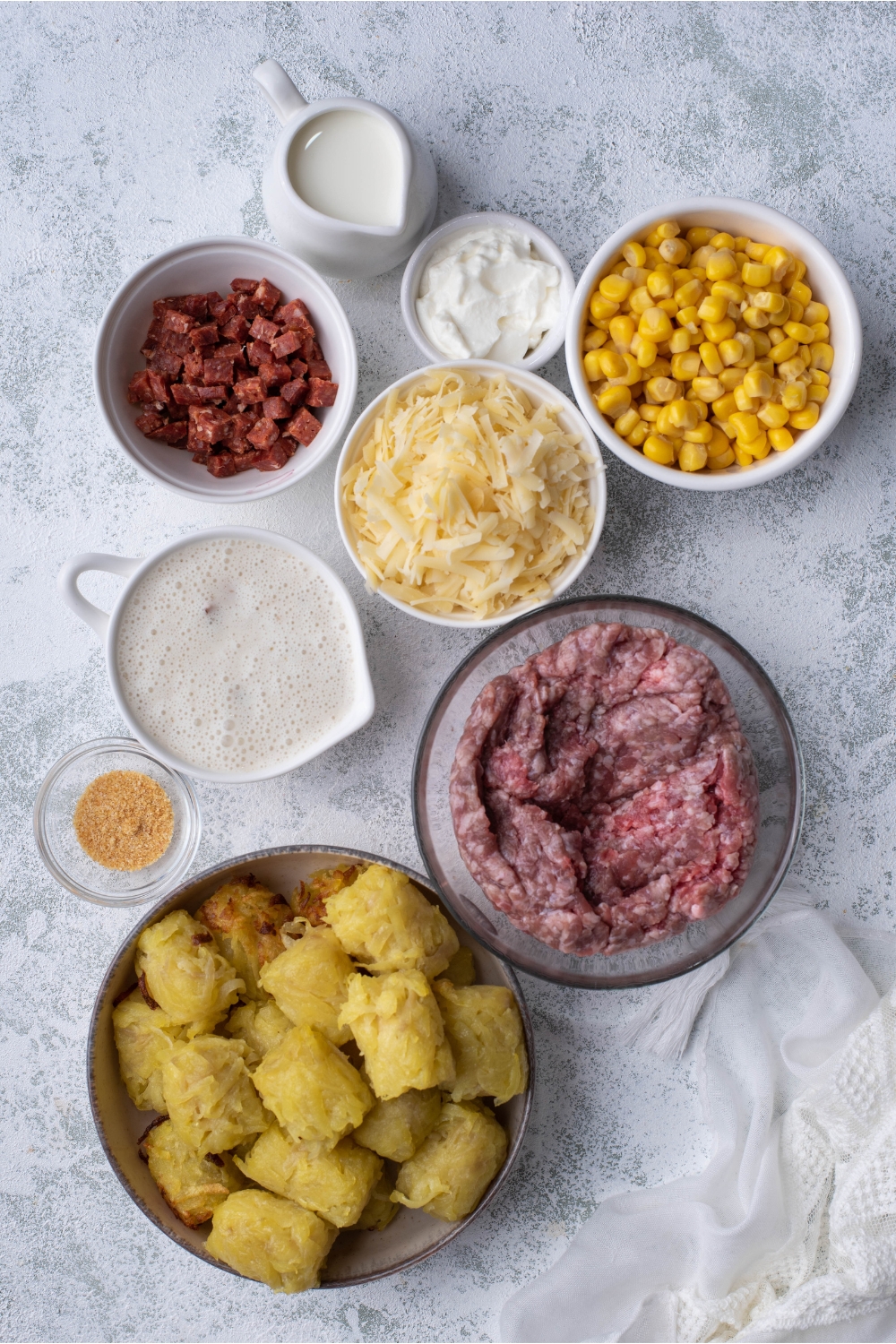 An assortment of ingredients including bowls of raw ground beef, corn, tater tots, chorizo, shredded cheese, sour cream, and spices.