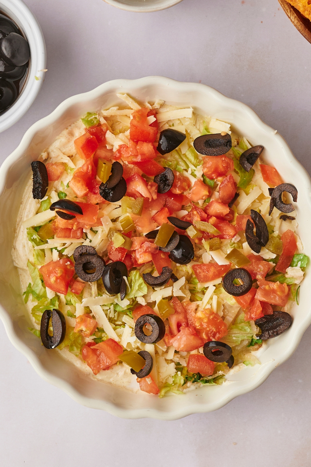 A serving bowl with creamy taco dip base, shredded lettuce, diced tomatoes, and sliced olives on it.