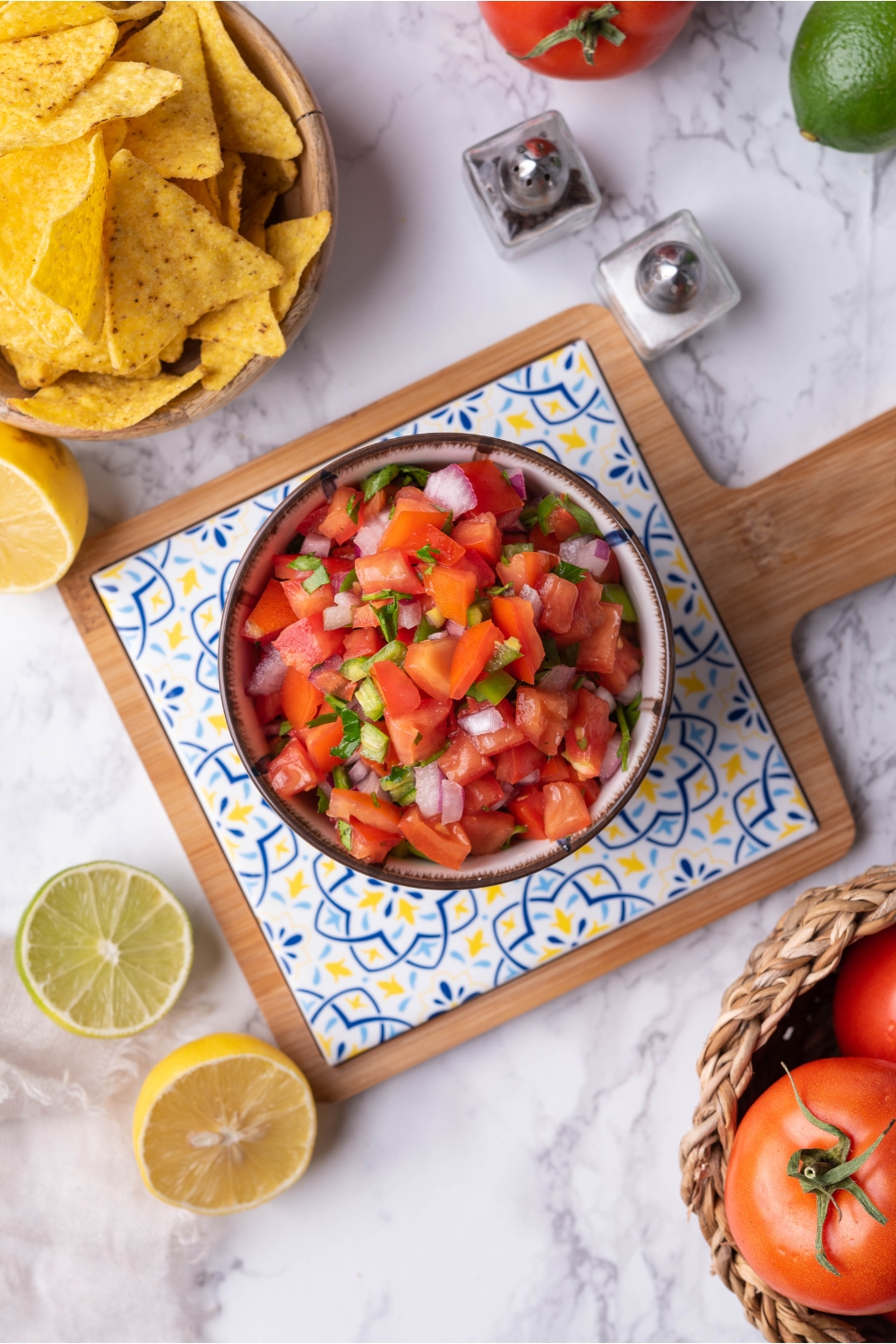 Overview of fresh tomato salsa on a decorative serving tray, surrounded by lemons, limes, tortilla chips, and salt and pepper shakers.