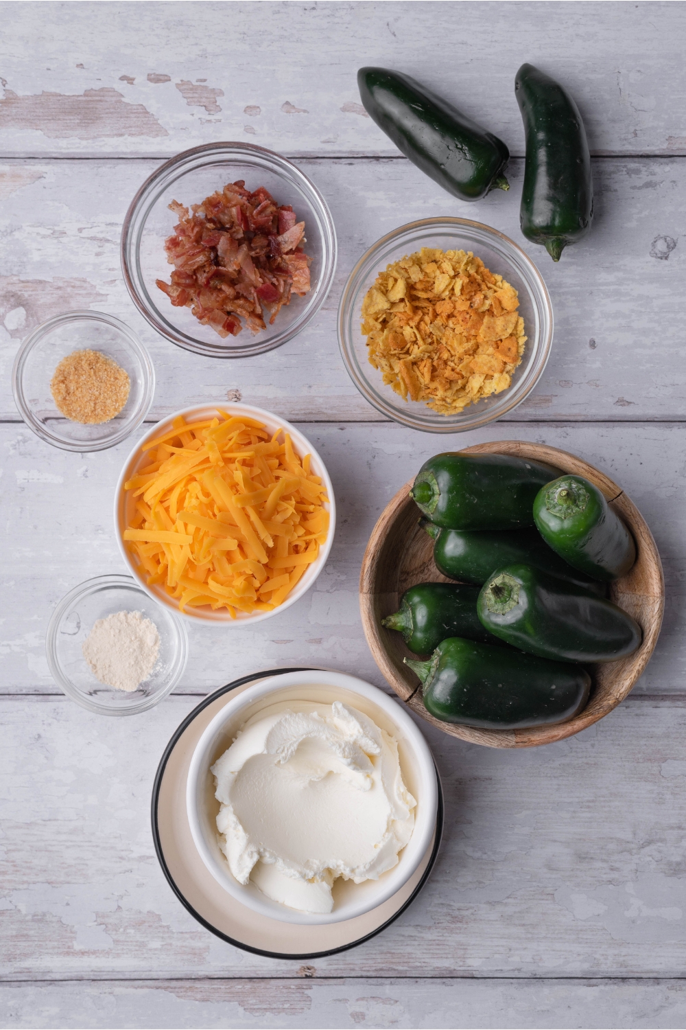 An assortment of ingredients including bowls of fresh jalapeños, cream cheese, shredded cheese, crumbled bacon, and crushed tortilla chips.