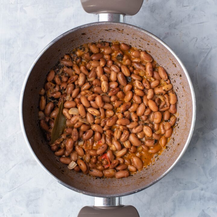 Grey sauce pot filled with cooked and seasoned pinto beans with a bay leaf and sliced peppers in the pot.