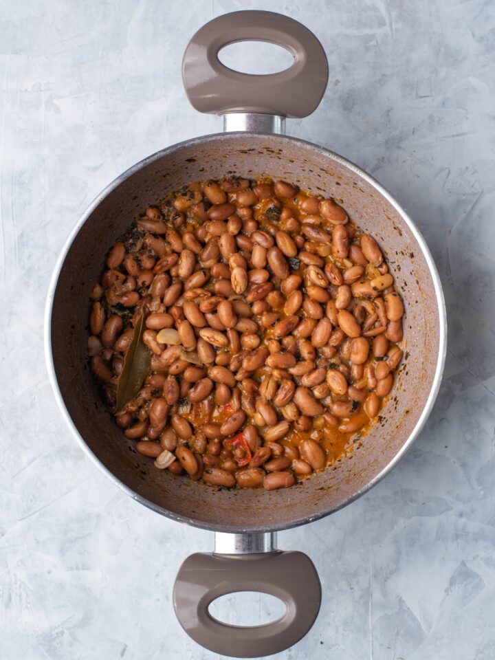 Grey sauce pot filled with cooked and seasoned pinto beans with a bay leaf and sliced peppers in the pot.