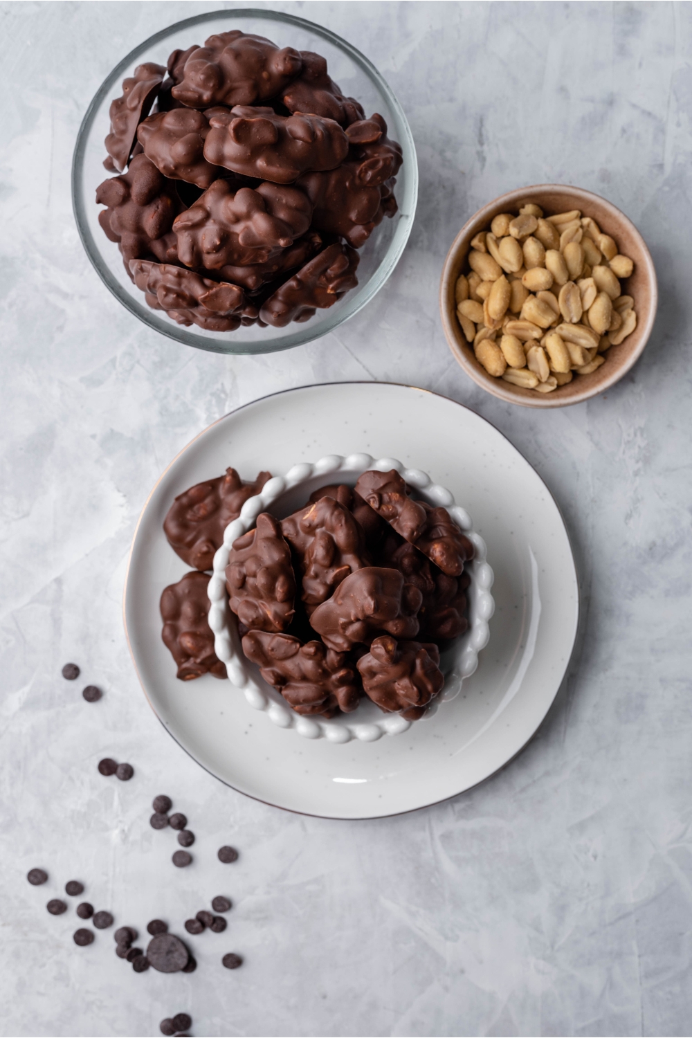 Overhead view of two bowls of chocolate peanut clusters and a bowl of peanuts. One bowl of clusters is atop a white plate with pieces falling over on the plate and chocolate chips are sprinkled on the counter.