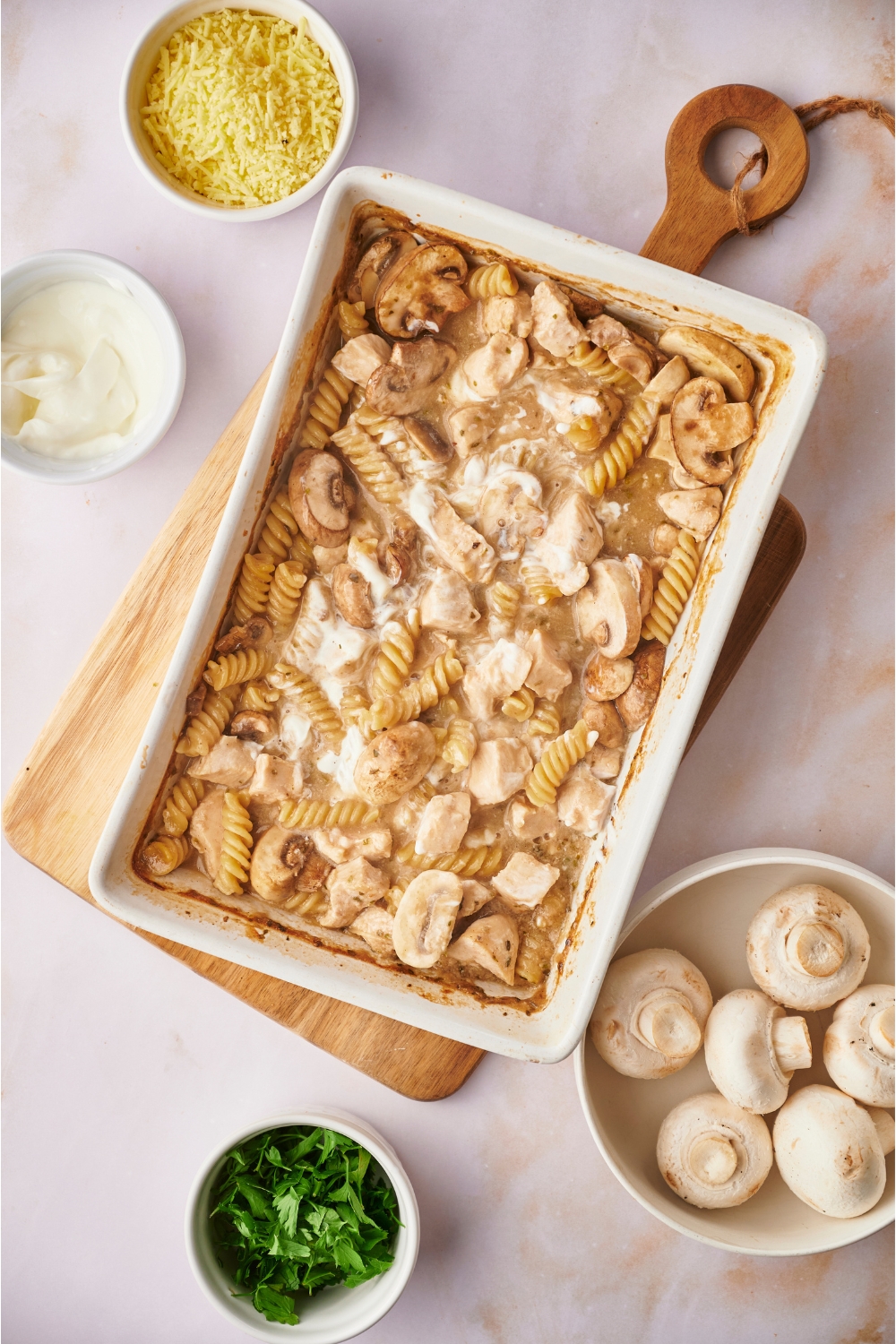 White baking dish on top of a wooden board filled with freshly baked chicken and mushroom casserole that has sour cream mixed on top. The dish is surrounded by ingredients including bowls of mushrooms, sour cream, and herbs.