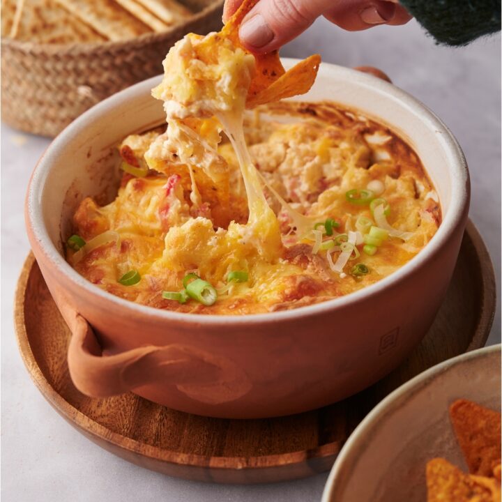 Hand scooping cheesy crab dip out of a baking dish with a tortilla chip.
