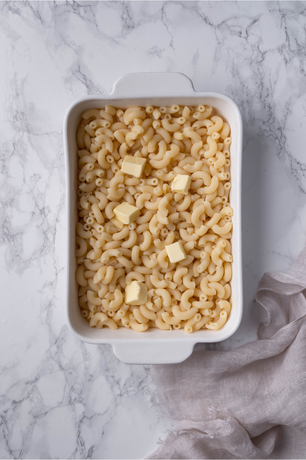 A white baking dish filled with cooked elbow macaroni and pats of butter atop the noodles.
