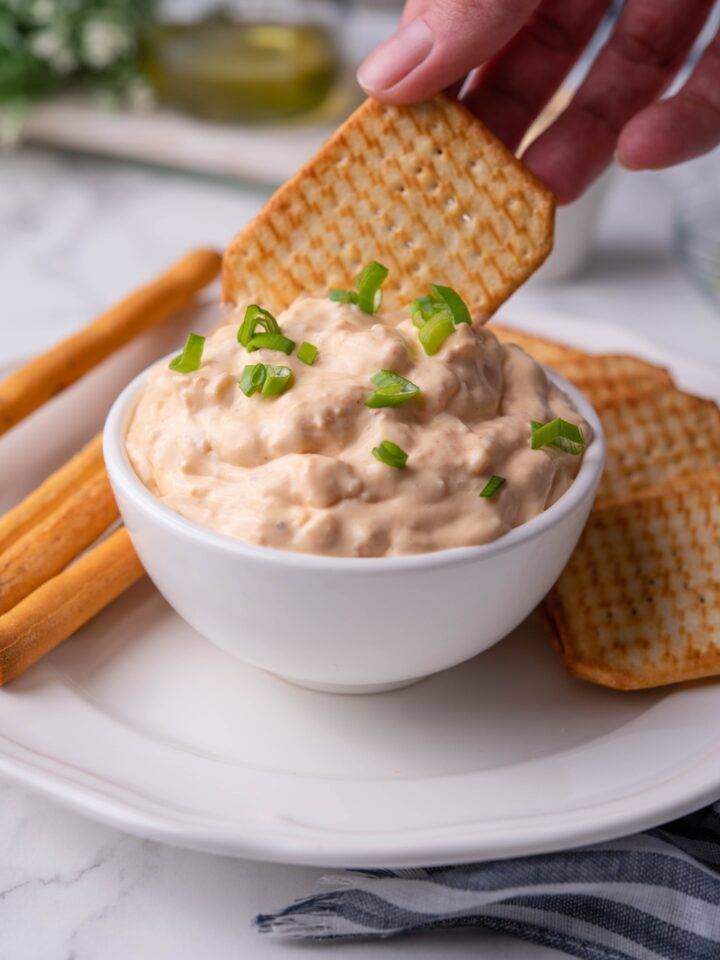 Hand dipping a cracker into a bowl of shrimp dip that's garnished with diced green onion.