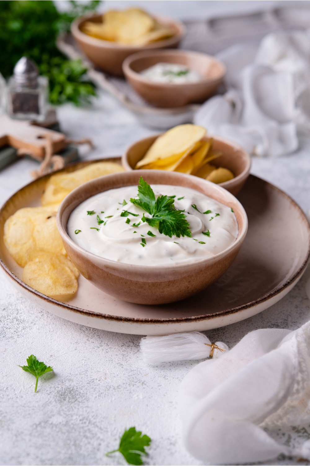 Sour cream dip garnished with fresh herbs in a brown bowl layered atop a brown plate. There are chips on the plate and in a small bowl next to the bowl of dip.