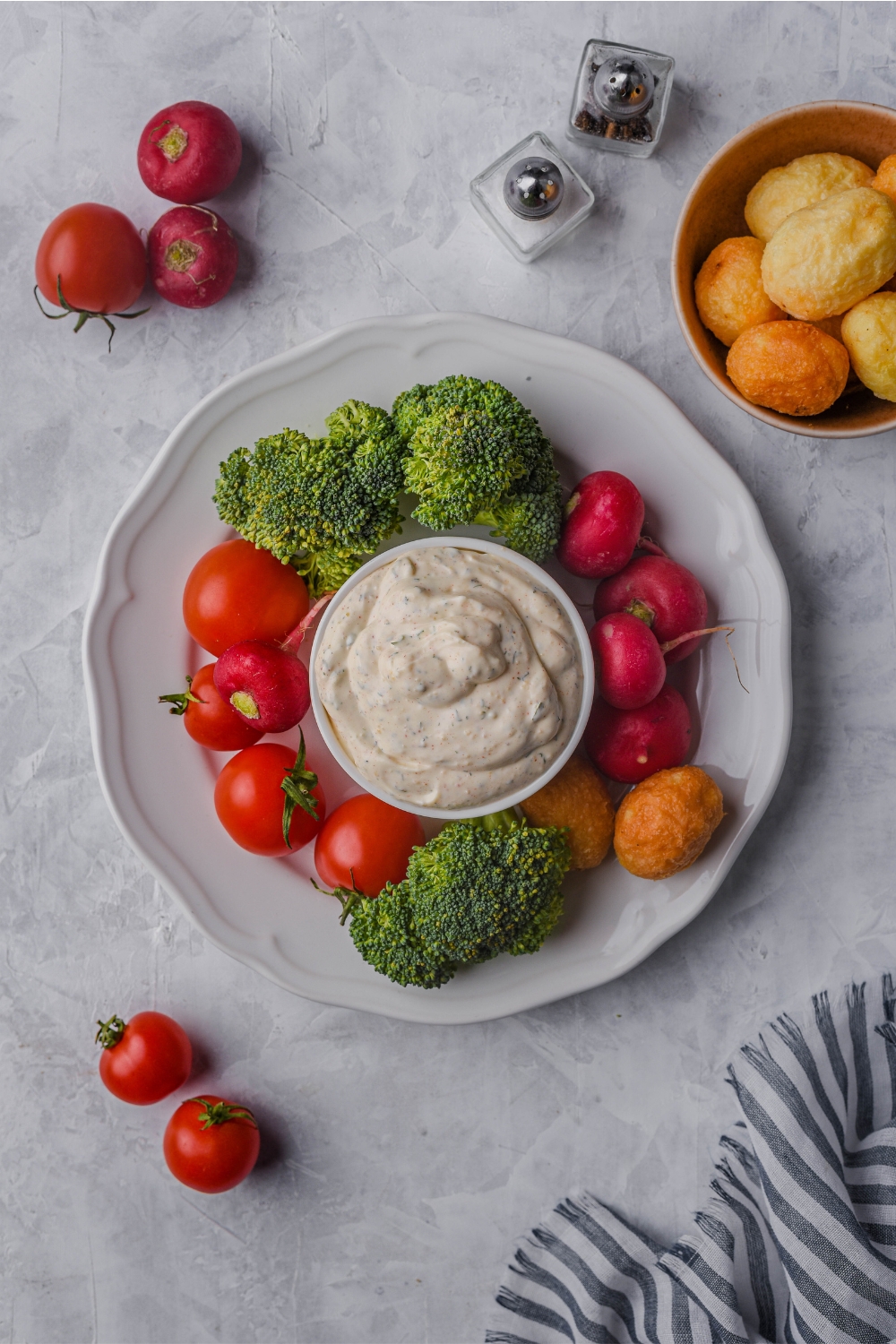 Overview of veggie dip in a white ramekin atop a white serving tray that is filled with broccoli, tomatoes, radishes, and nuggets. Surrounding the plate are cherry tomatoes, radishes, salt and pepper shakers, and a bowl of nuggets.