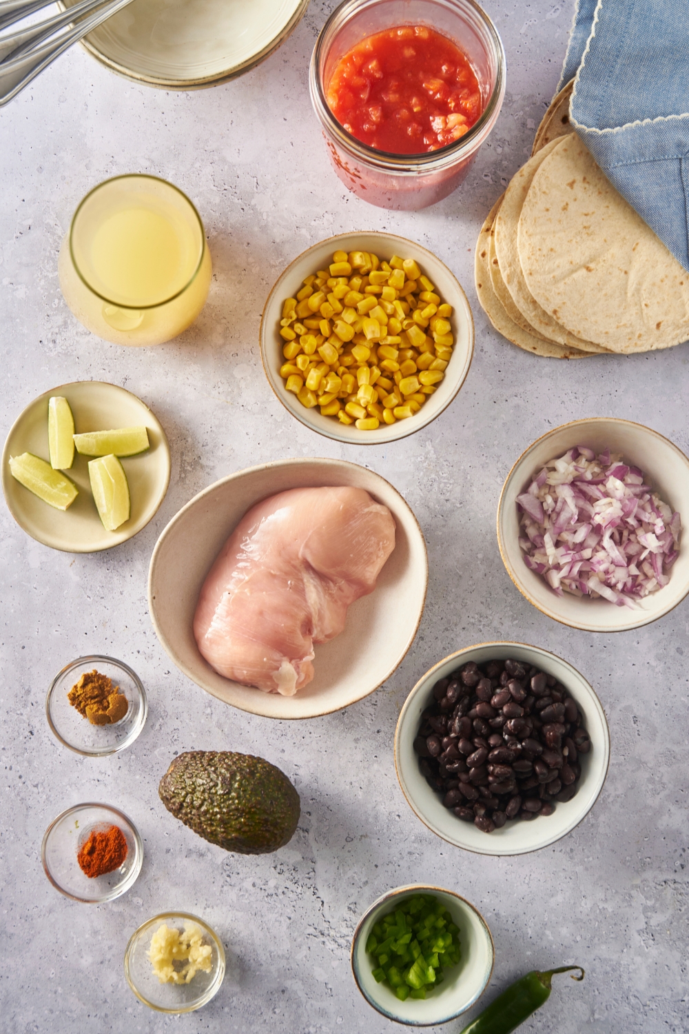 An assortment of ingredients including bowls of raw chicken, diced red onion, corn, peppers, spices, lime wedges, and black beans.
