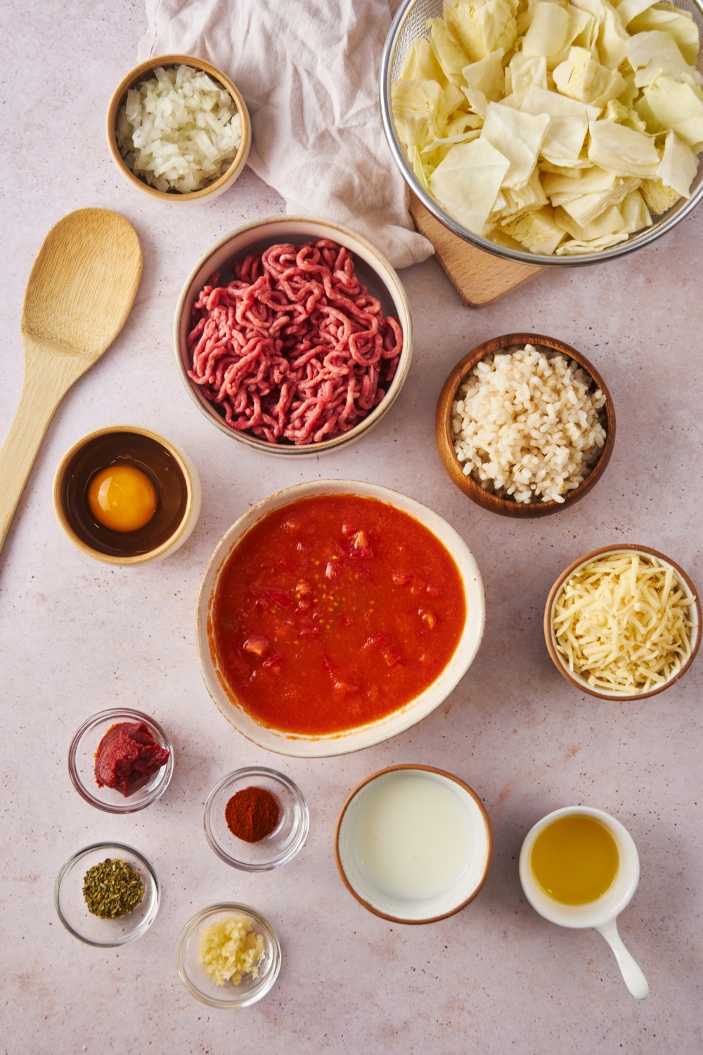 An assortment of ingredients including bowls of raw beef, sliced cabbage, tomato sauce, shredded cheese, milk, and spices.