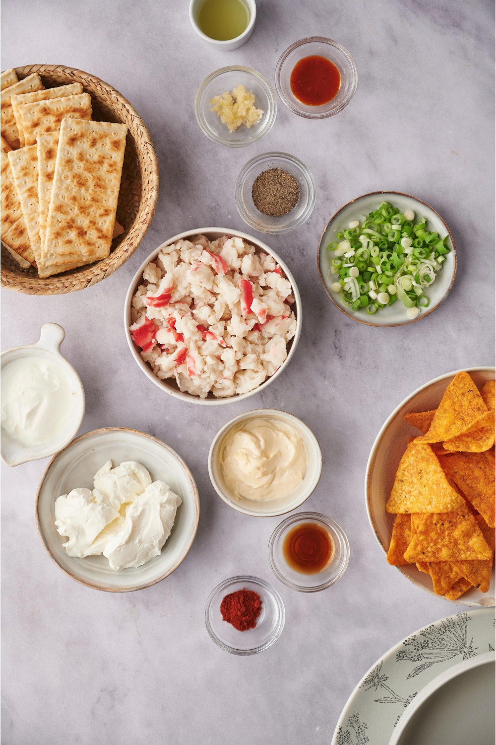 An assortment of ingredients including bowls of chopped crab, diced green onion, mayonnaise, sour cream, cream cheese, spices, crackers, and tortilla chips.