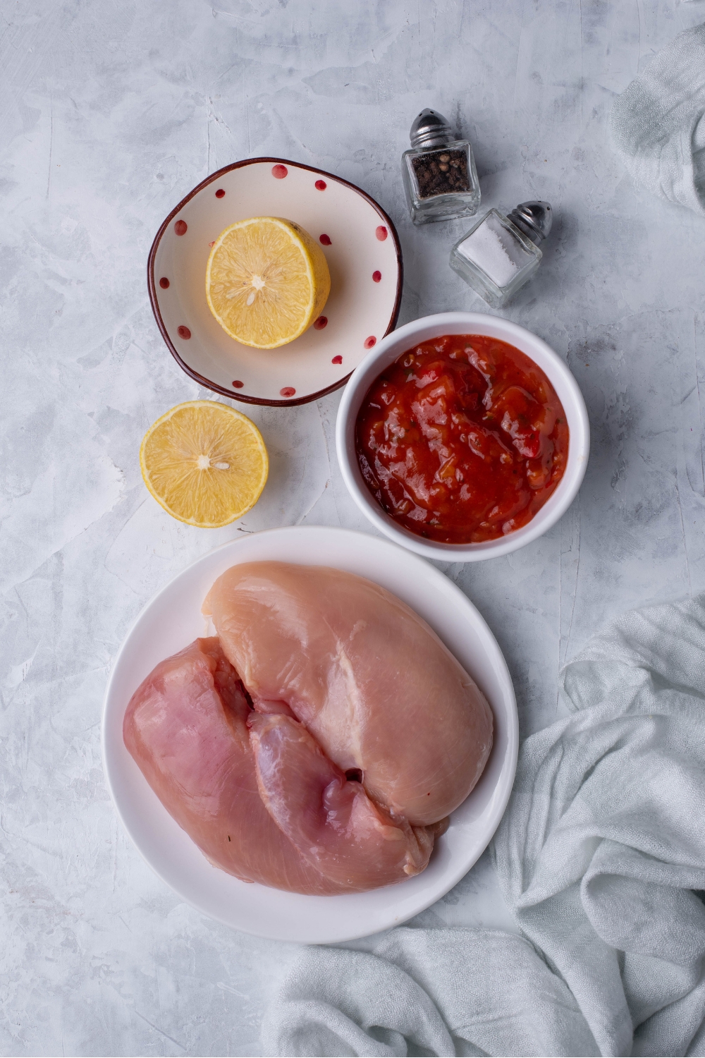 An assortment of ingredients including a plate of raw chicken breast, a bowl of salsa, salt and pepper shakers, and a lemon cut in half with one half in a bowl and one half on the counter.