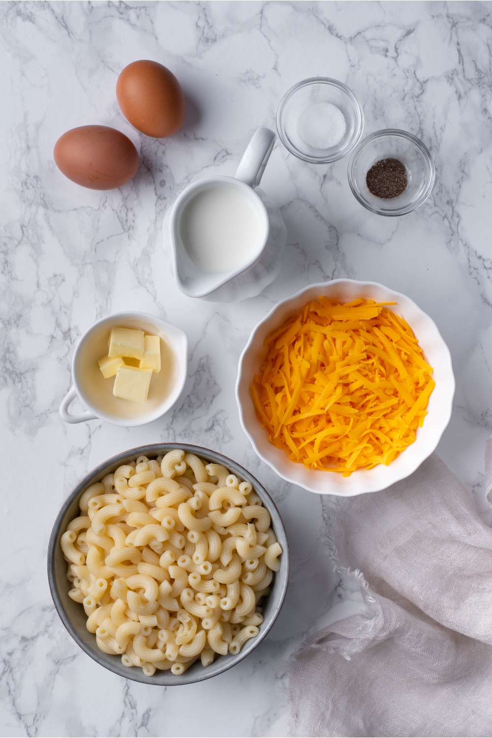 An assortment of ingredients including bowls of shredded cheese, elbow macaroni, butter cubes, salt, pepper, a container of milk and two eggs.