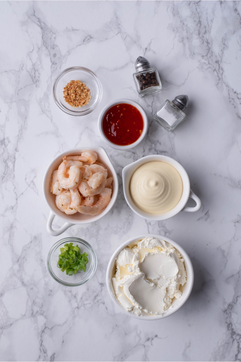 An assortment of ingredients including bowls of cooked shrimp, mayonnaise, cream cheese, diced green onion, chili sauce, and spices.