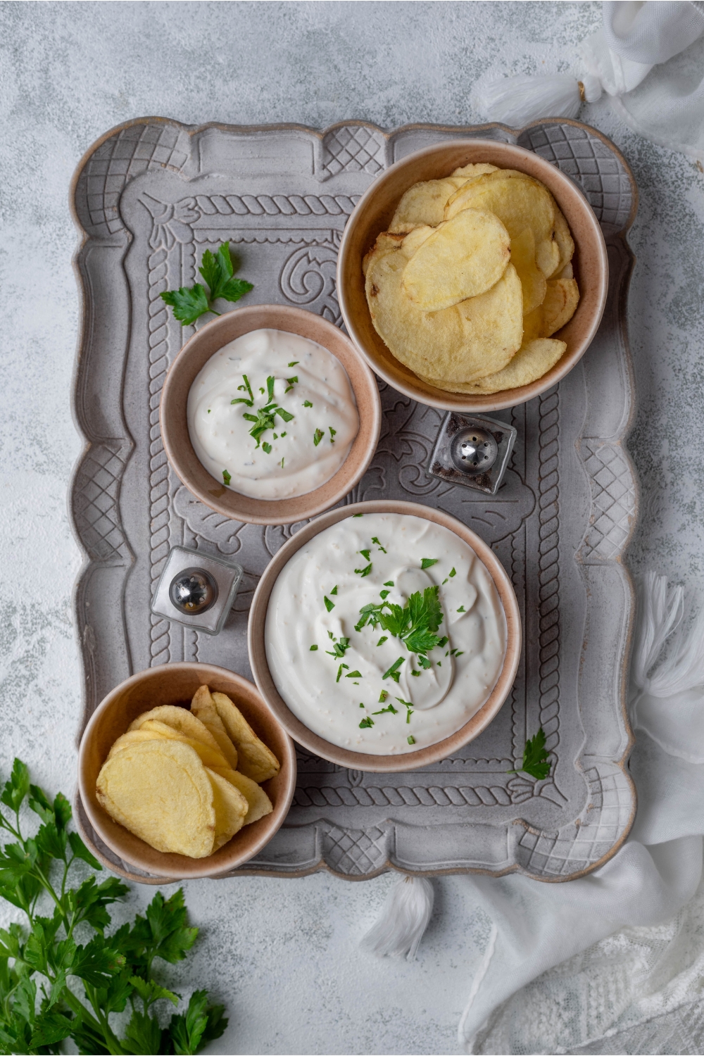 Overview of two bowls of sour cream dip and two bowls of chips on a decorative grey tray. There's fresh herbs garnished on the dip and sprinkled on the tray alongside a set of salt and pepper shakers.