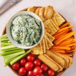 An overhead view of a serving bowl with spinach dip on a tray with vegetables, crackers, and bread surrounding it.