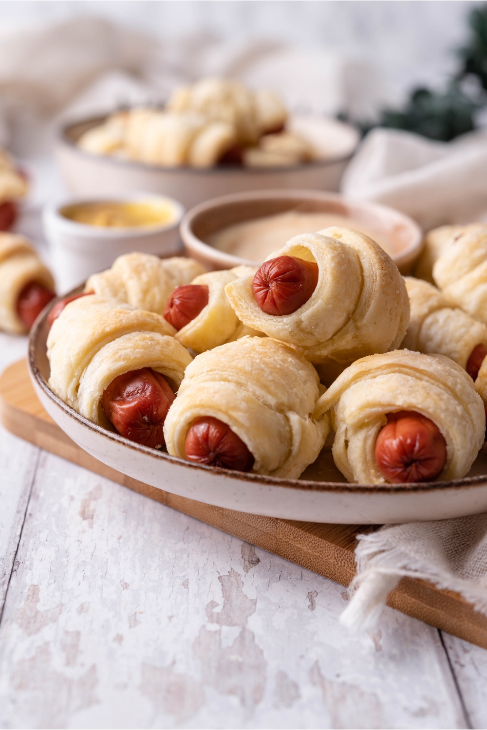 Pigs in a blanket piled on top of each other along with a bowl of spicy mayo on a white plate atop a wood board.