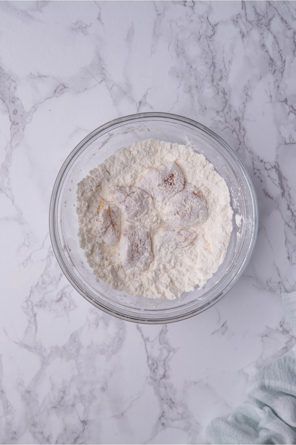 Clear bowl filled with raw chicken pieces in a flour mixture.