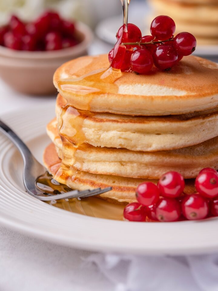 A stack of pancakes topped with fresh berries. Fresh syrup is being poured over the stack.