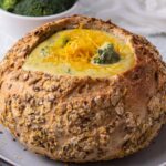 A bread bowl with broccoli cheese soup topped with extra cheddar cheese.