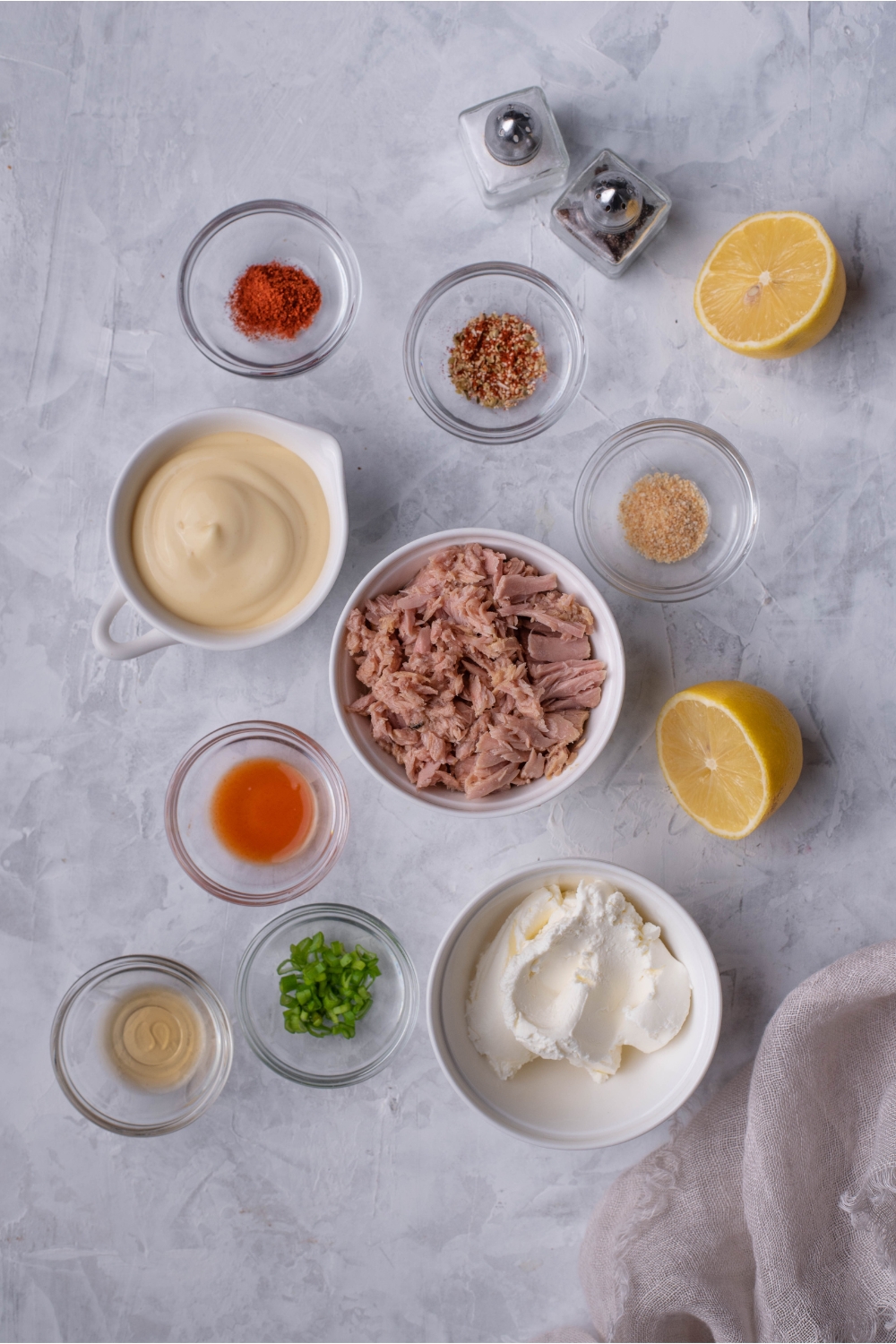 An assortment of ingredients including bowls of mayo, canned tuna, cream cheese, spices, two lemon halves, and salt and pepper shakers.
