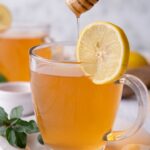 A clear mug with medicine ball tea in it, a slice of lemon is garnished on the side. A honey stick is drizzling honey into the tea.