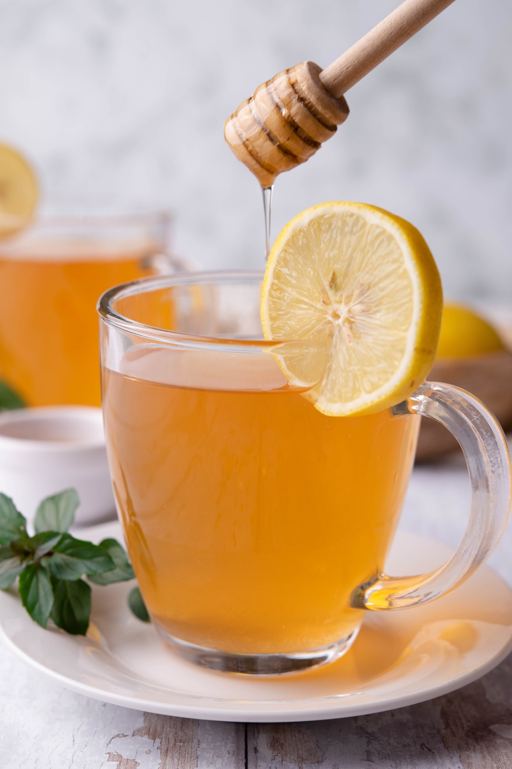 A clear mug with medicine ball tea in it, a slice of lemon is garnished on the side. A honey stick is drizzling honey into the tea.