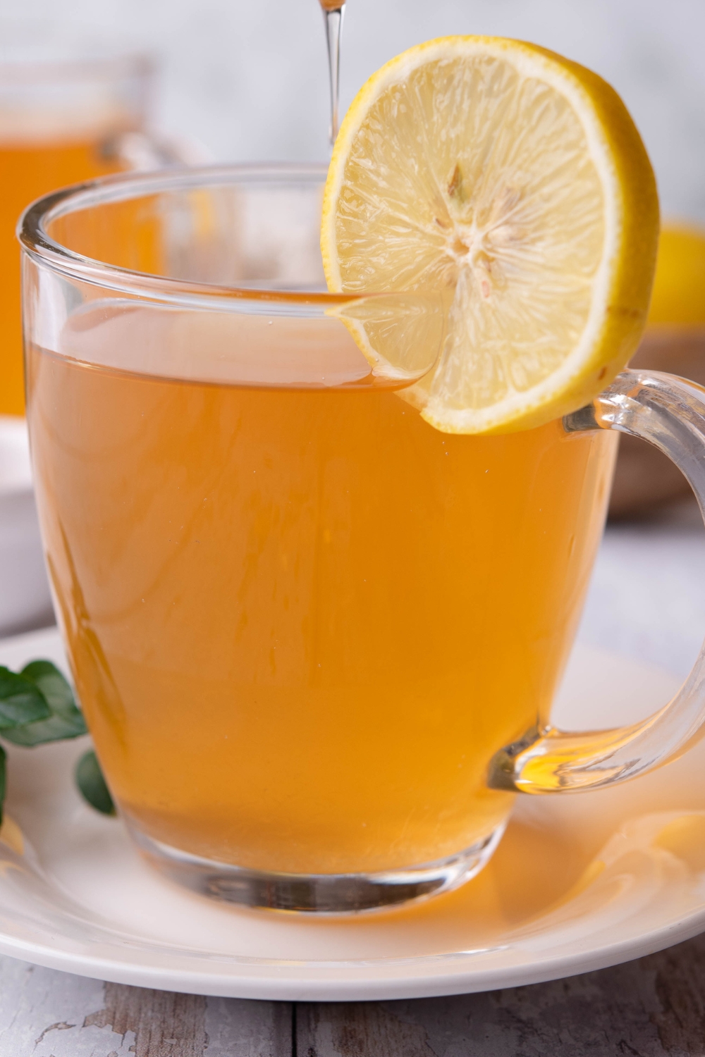 A clear mug with medicine ball tea with a slice of lemon garnishing it and honey being drizzled into it.