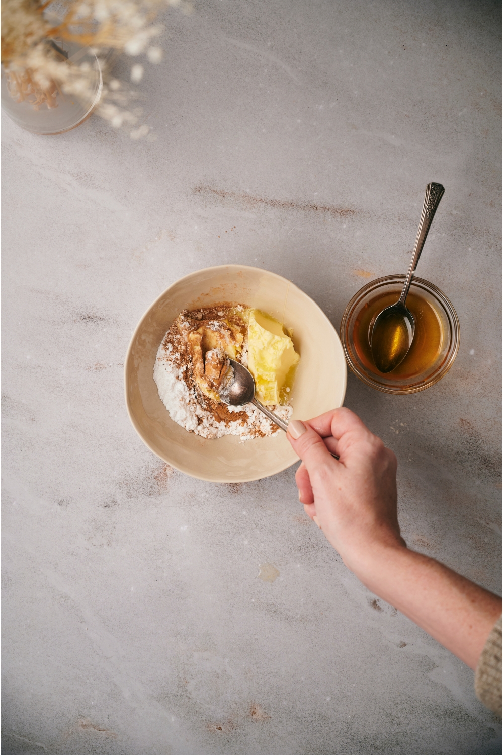A hand mixing butter, cinnamon, honey, and powdered sugar together with a spoon, next to a small bowl of honey.