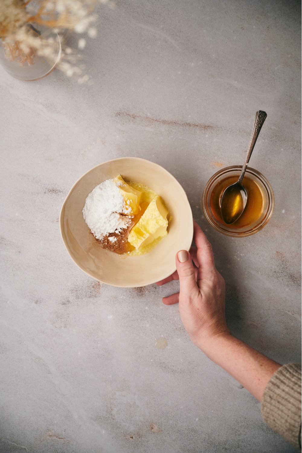 An off-white bowl filled with softened butter, powdered sugar, cinnamon, and honey, with a hand positioning the bowl next to a small bowl of honey with a spoon in it.