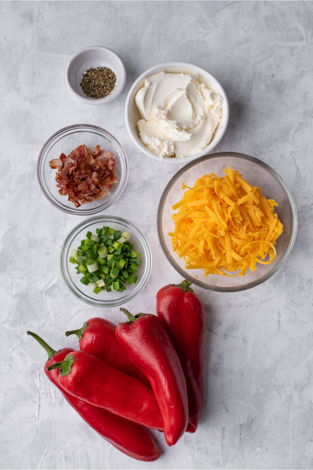 An assortment of ingredients including a pile of sweet red peppers and bowls of cream cheese, shredded cheese, bacon, sliced green onion, and black pepper.