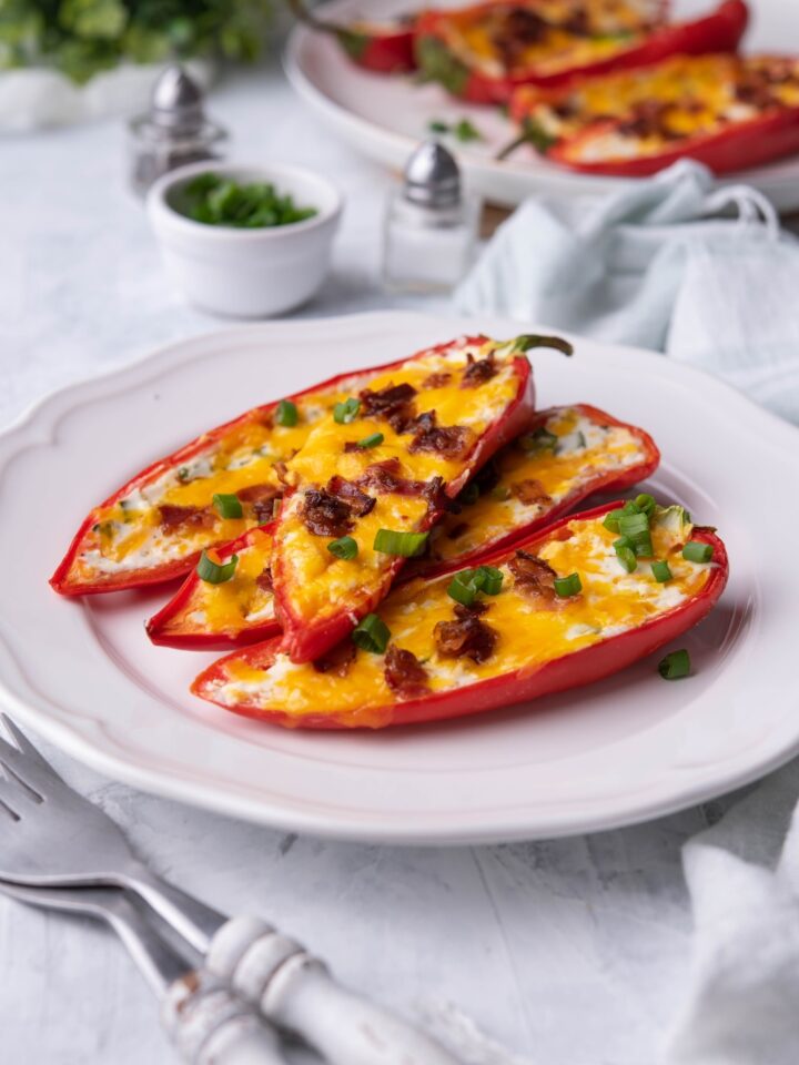 Four cream cheese stuffed peppers piled on top of each other on a white plate next to two forks with a second plate of stuffed peppers in the background.
