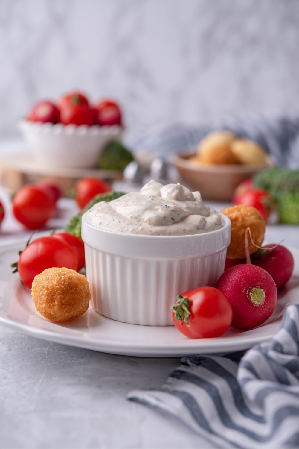 Vegetable dip in a white ramekin atop a white plate that is filled with cherry tomatoes and nuggets. In the background are bowls of tomatoes, nuggets, and fresh vegetables.