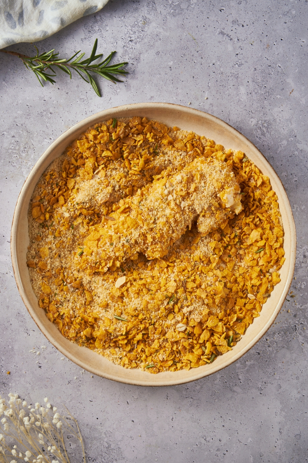 Breaded chicken tenders in a bowl filled with a mixture of cornflakes, bread crumbs and spices.