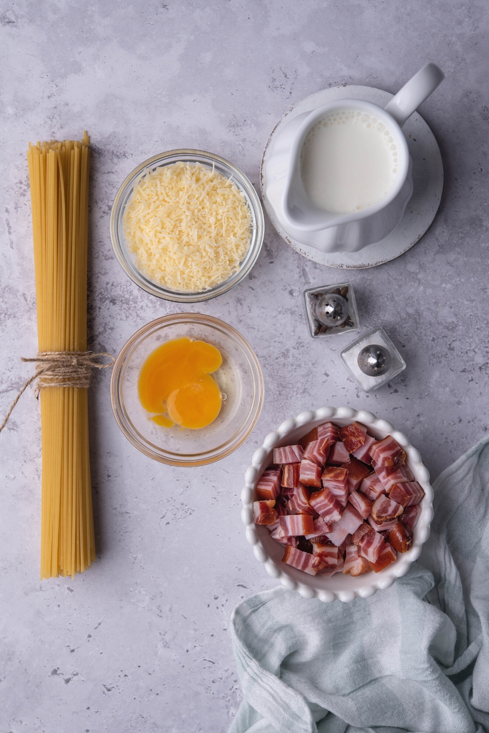 A bowl of pancetta, a bowl of egg yolks, a bowl of grated Parmesan cheese, a bundle of pasta noodles, and a picture of cream all on a gray counter.