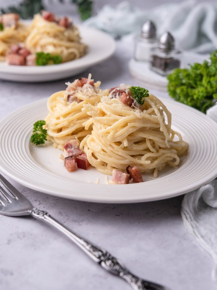 Two stacks of spaghetti noodles with diced pancetta and Parmesan cheese on them on a white plate.