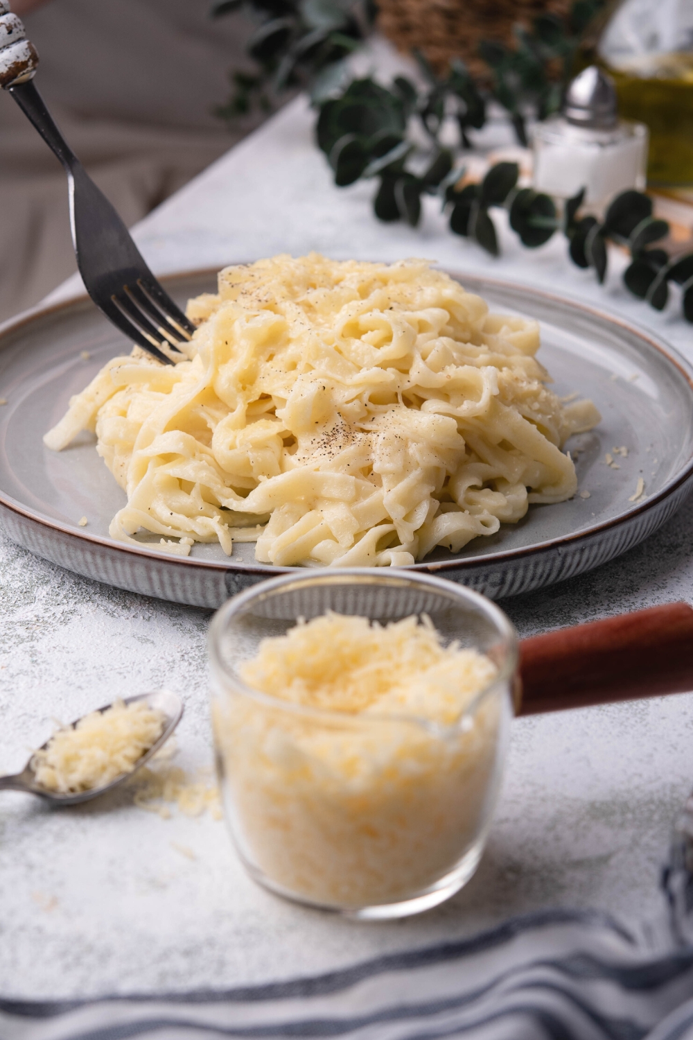 A fork being submerged in a pile of parmesan noodles on a plate. There is a cup of parmesan cheese in front of it.