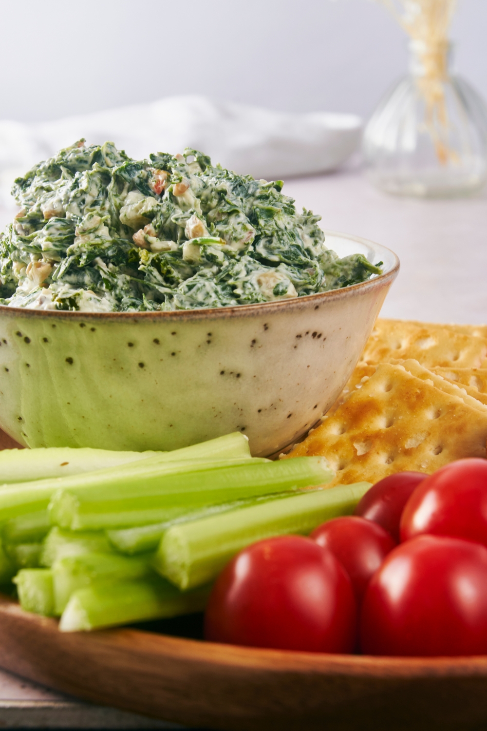 A serving bowl with spinach dip on a tray with vegetables, crackers, and bread surrounding it.