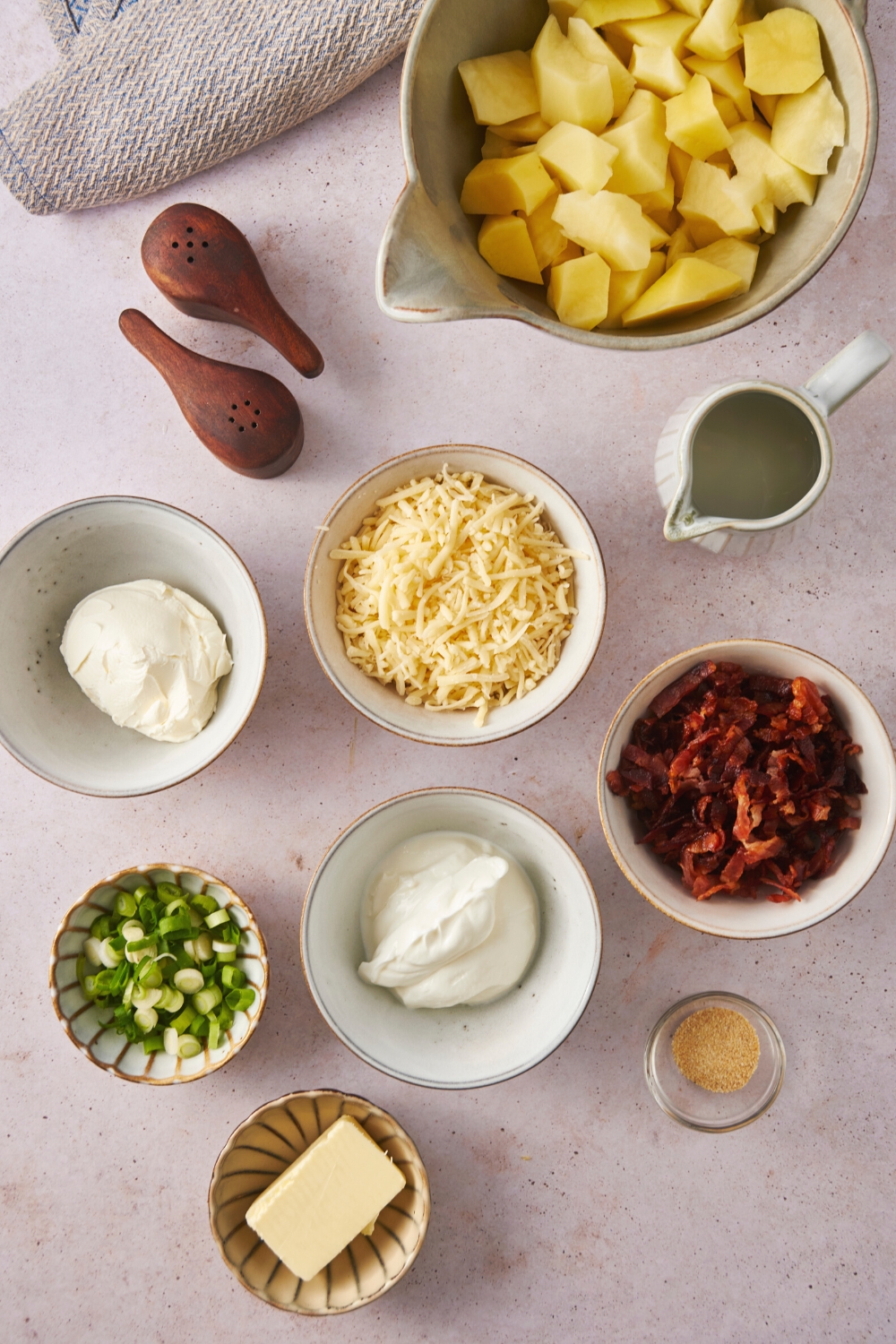 A bowl of diced potatoes, a bowl of cream cheese, a bowl of shredded cheese, a bowl of bacon bits, a bowl of chopped green onion, a bowl of sour cream, and a bowl of butter all on a counter.