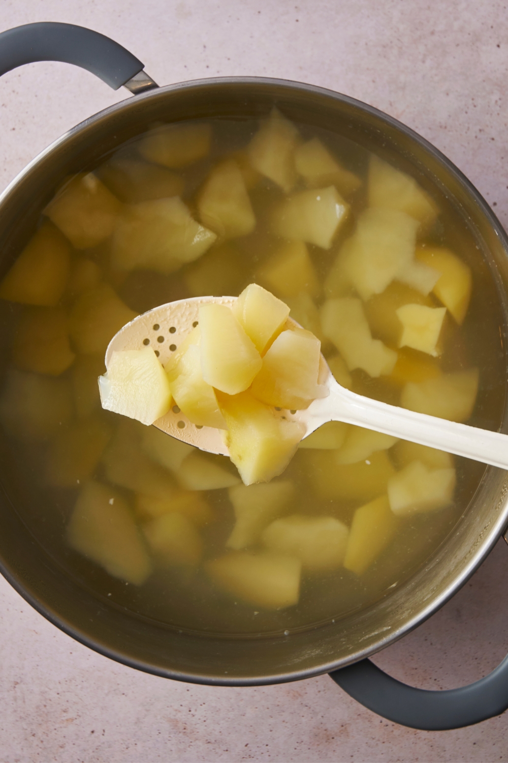A slotted spoon holding diced potatoes over a deep pot with the remaining cubed potatoes in hot water.