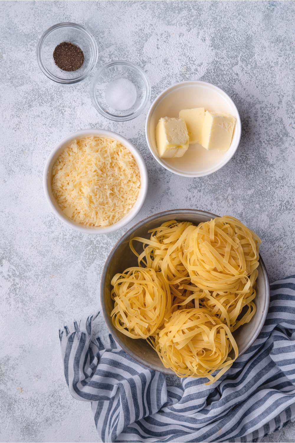 A bowl of pepper, a bowl of salt, a bowl of three cubes of butter, a bowl of parmesan cheese, and a bowl of three bundles of pasta.