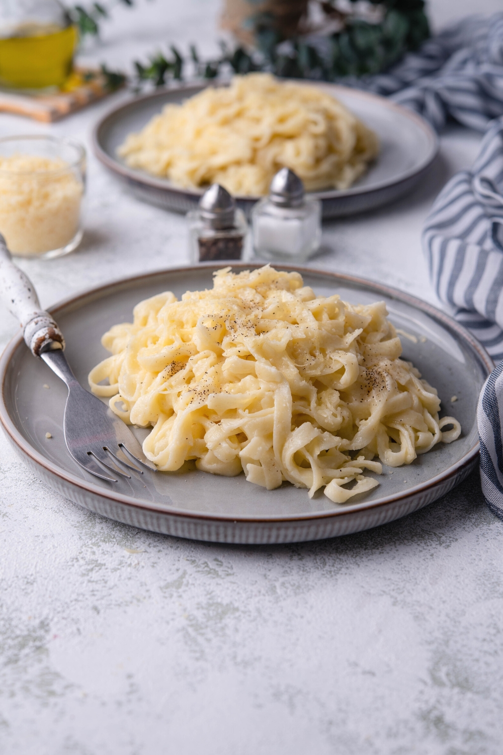 A pile of parmesan noodles on a plate with a fork on it. Behind it is a salt and pepper shaker and another plate of noodles.