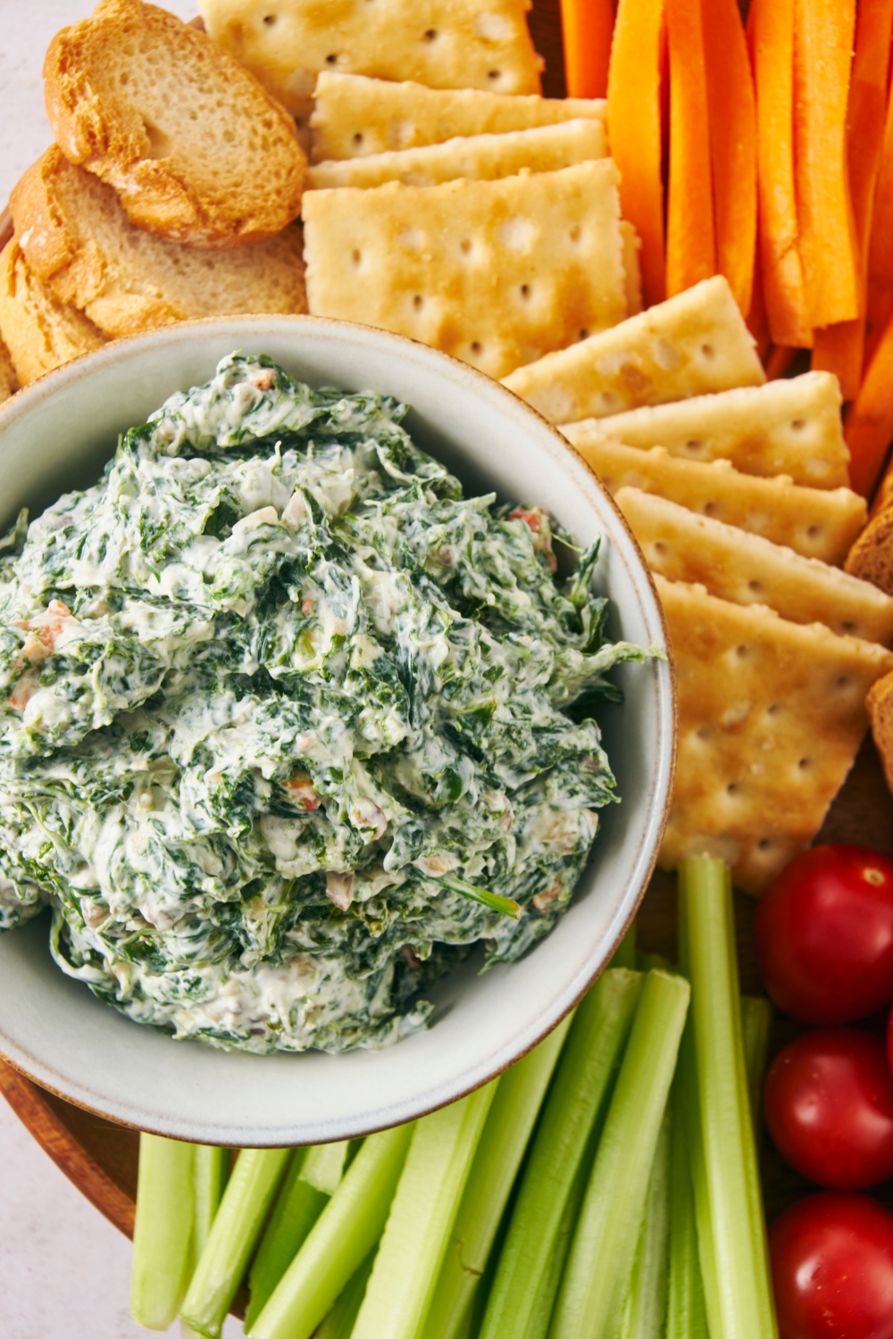 An overhead view of a serving bowl with spinach dip on a tray with vegetables, crackers, and bread surrounding it.