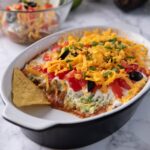 7 Layer dip in a black and white baking dish with a serving scooped out. There is a chip in the baking dish and more chips on the counter.