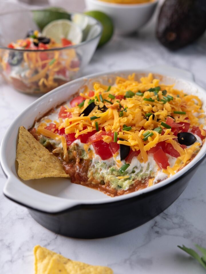 7 Layer dip in a black and white baking dish with a serving scooped out. There is a chip in the baking dish and more chips on the counter.