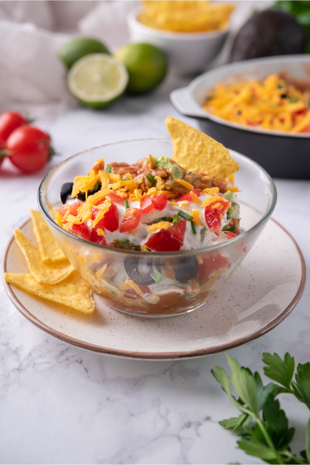 7 layer dip in a clear bowl atop a brown-rimmed plate, with one tortilla chip in the bowl and three more chips on the plate. In the background is the rest of the dip, cherry tomatoes, limes, and a bowl of tortilla chips.